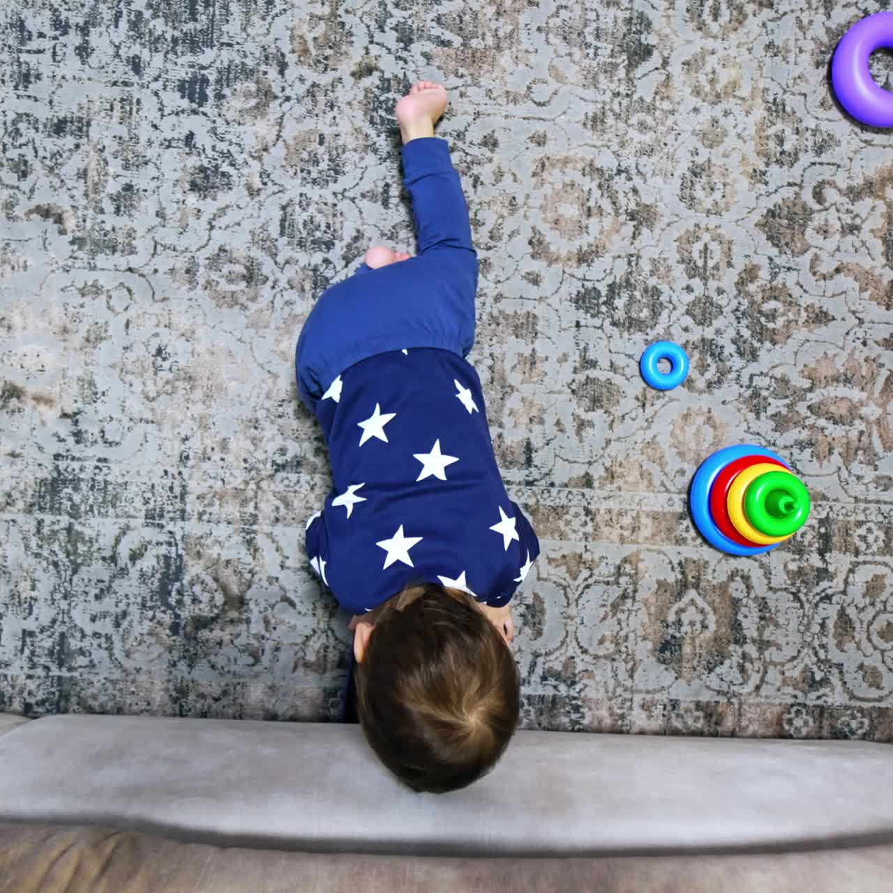 Little baby crawling by the carpet. Kid reaches his hand under the sofa and pulls the phone out. Child playing with gadget. Top view