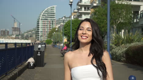 Slow-Motion-of-an-attractive-and-playful-latina-woman-with-black-wavy-hair-walking-on-the-banks-of-the-Thames-river-in-London,-looking-at-the-camera,-spinning,-happy-with-a-beautiful-smile