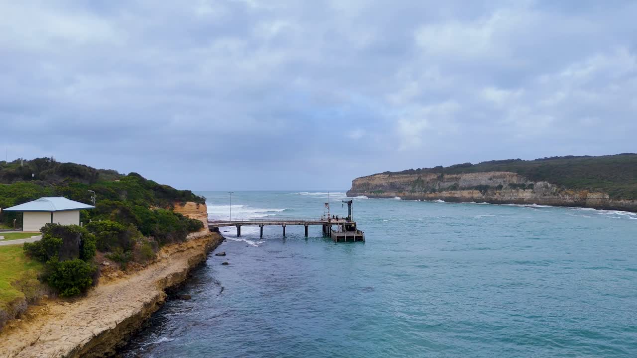 Aerial view of Port Campbell's pier and bay, showcasing coastal erosion and ocean waves under cloudy skies. Captured with smooth drone movement