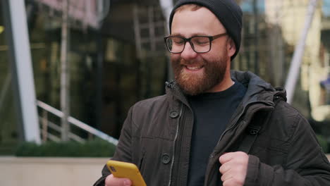 happy bearded man in beanie and glasses using smartphone outdoors