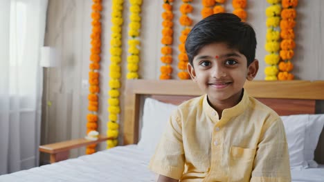 indian boy smiling sitting on bed in decorated room with orange and yellow flower garlands hanging on the wall for a traditional hindu celebration