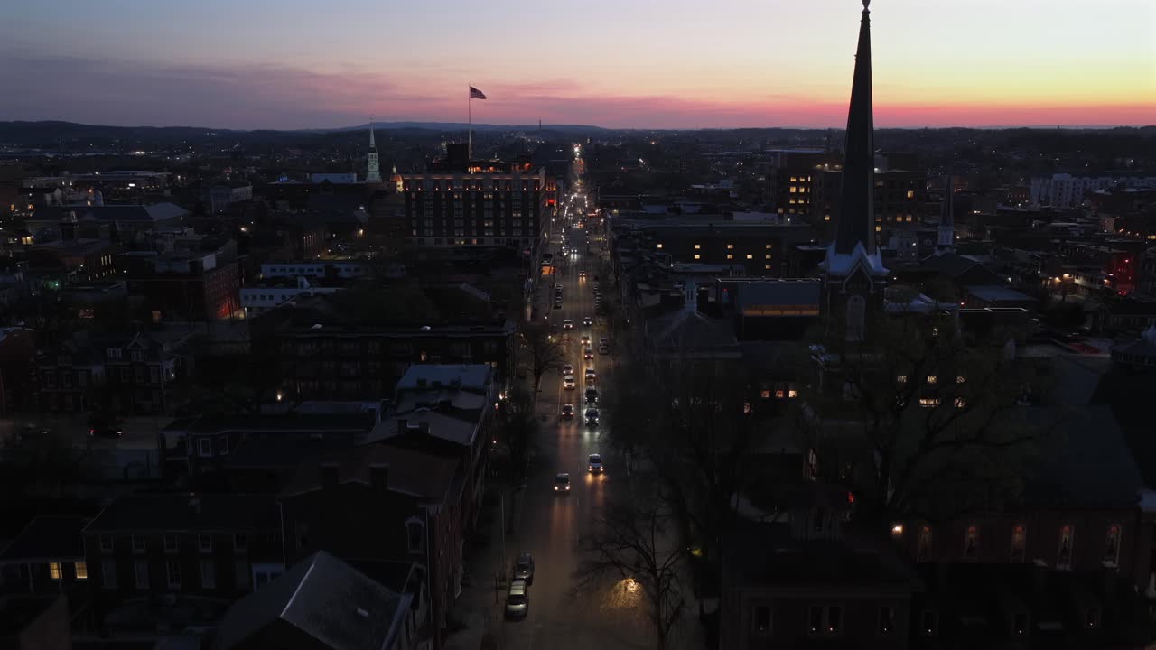 Elevación aérea sobre el centro de York, Pensilvania, al atardecer, con tráfico iluminado, un campanario de iglesia, la bandera estadounidense y tejados históricos bajo un cielo crepuscular colorido en el centro de la ciudad. Toma panorámica. Escena de tráfico.