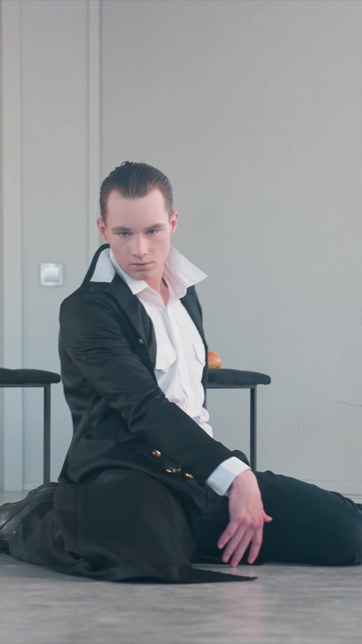 Man in black coat and white shirt performs dramatic gestures. Man interacts with his hands above head chest stomach hip. Indoor setting features light colored walls minimal