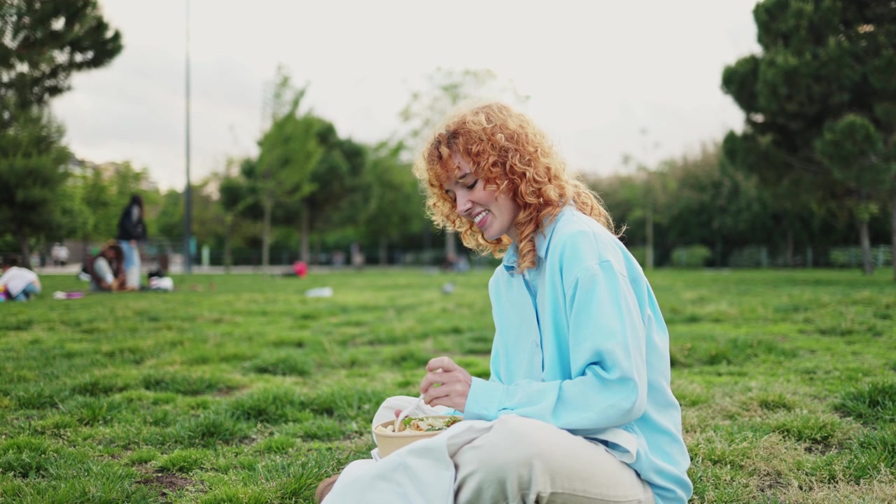 Glad ung kvinde med krøllet hår spiser salat i parken