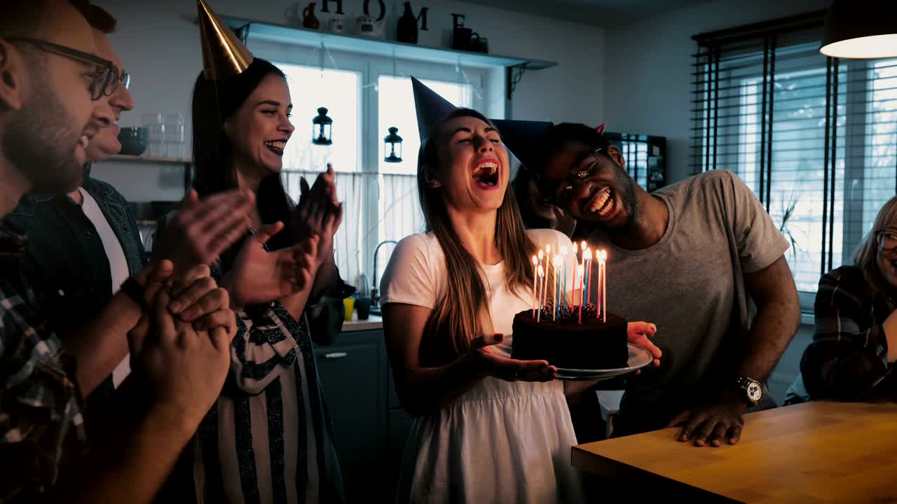 Happy European girl holding birthday cake, making a wish and blowing, multiethnic friends sing at fun party slow motion