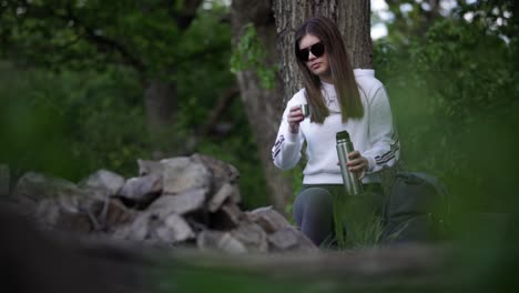 joven caucásica sentada en el bosque, sirviéndose una bebida caliente de una botella termo y bebiéndola