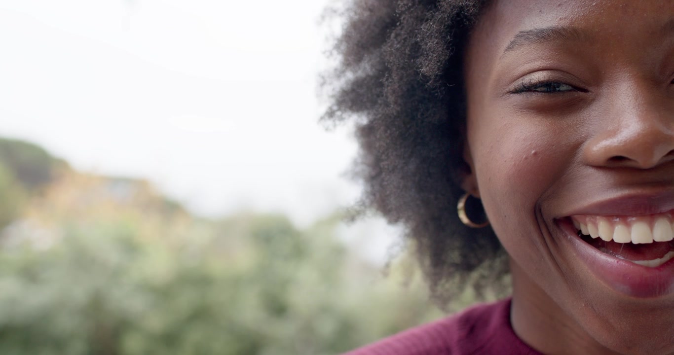 Adult African woman hearing laugh, shifting from smile to laugh in garden, maroon-top, copy space