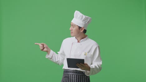 chef asiático usando una tableta y señalando hacia el lado mientras está de pie en el estudio de fondo de la pantalla verde