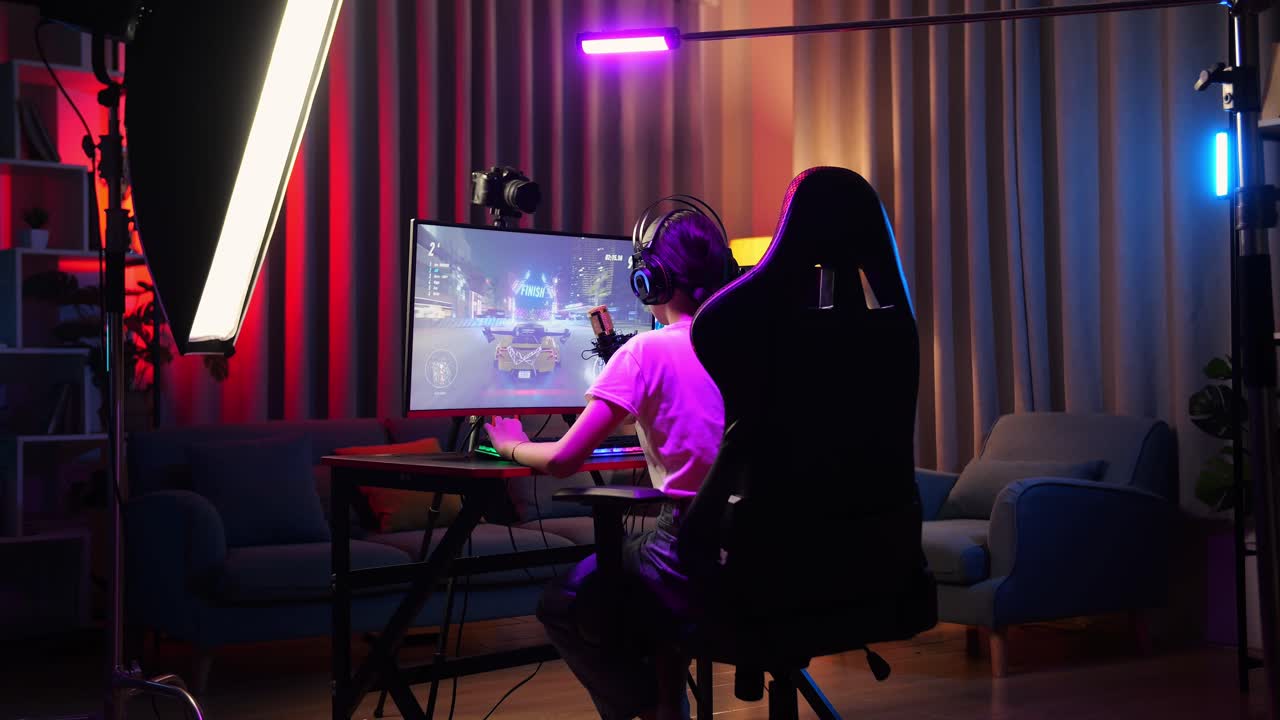 Vista trasera de una streamer asiática decepcionada tras perder un juego de carreras de coches en su ordenador personal. Transmisión en vivo de un videojuego, escritorio iluminado por una tira de luz LED RGB.