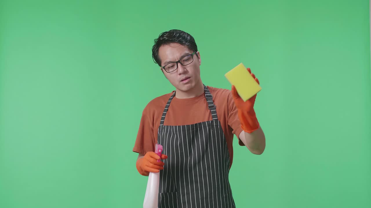 cansado ama de casa masculina asiática con un delantal sosteniendo un spray y una esponja de cocina luego limpiando el sudor en el estudio de pantalla verde