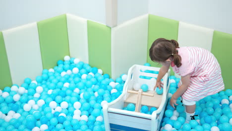 Una-Niña-De-3-Años-Juega-En-Una-Piscina-Seca-Con-Bolas-De-Plástico-Blancas-Y-Azules,-Lanza-Bolas-En-Un-Barco-De-Juguete-De-Madera