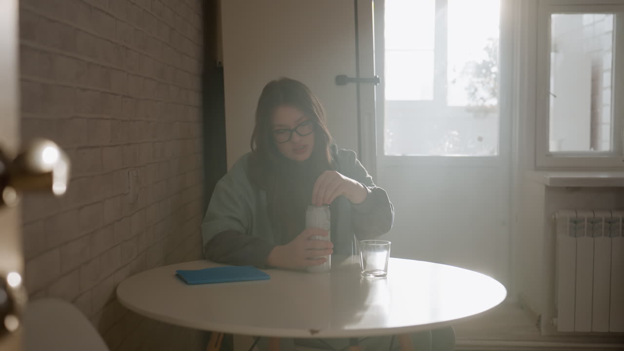 Donna al tavolo della cucina illuminato dal sole che versa il latte in un bicchiere, accogliente scena mattutina con una studentessa con gli occhiali vicino alla finestra, calda retroilluminazione e ombre soffuse, arredamento minimale, atmosfera tranquilla e riflessiva.