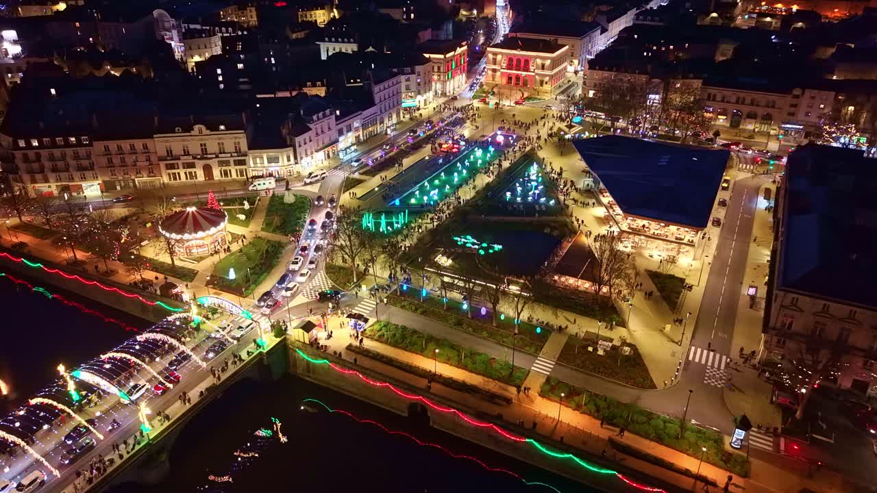 Forward drone shot from the Mayenne toward Laval’s November 11 Square, showing glowing Christmas decorations, luminous animals, the carousel, the market hall and the decorated Aristide Briand Bridge