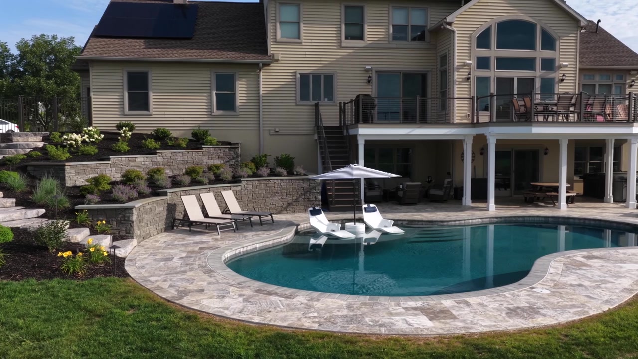 Curved pool reflects large home with solar panels and covered patio, white lounge chairs rest on tanning ledge beneath striped umbrella, stone terraces with flowers line landscaped backyard. Aerial