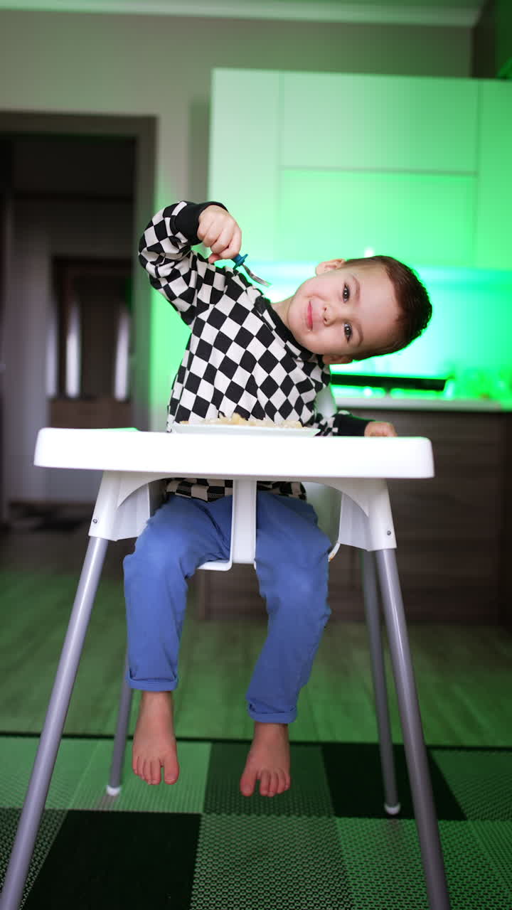 Lovely toddler sitting at high chair in the kitchen. Kid picks some food with his fork but drops it on the floor. Vertical video.