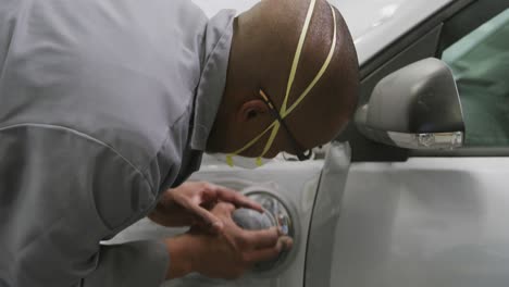 african american male car mechanic wearing a face mask and polishing a side of a car with a grinder