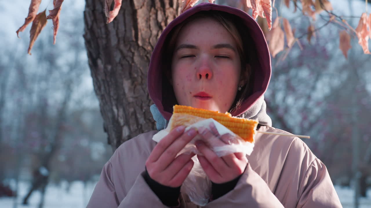 겨울 옷을 입은 젊은 여성이 나무에 앉아 옥수수를 먹고, 야외 공원에서 겨울 동안 간식, 배경에서 부드러운 빛 반사, 평온하고 조용한 분위기를 만드는, 겨울 공원 즐거움
