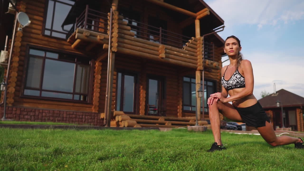 una mujer delgada y hermosa en ropa deportiva con una prensa abierta se está preparando para comenzar a entrenar en el césped cerca de su casa flexionando sus piernas, rodillas, espinillas y muslos con ejercicios de calentamiento