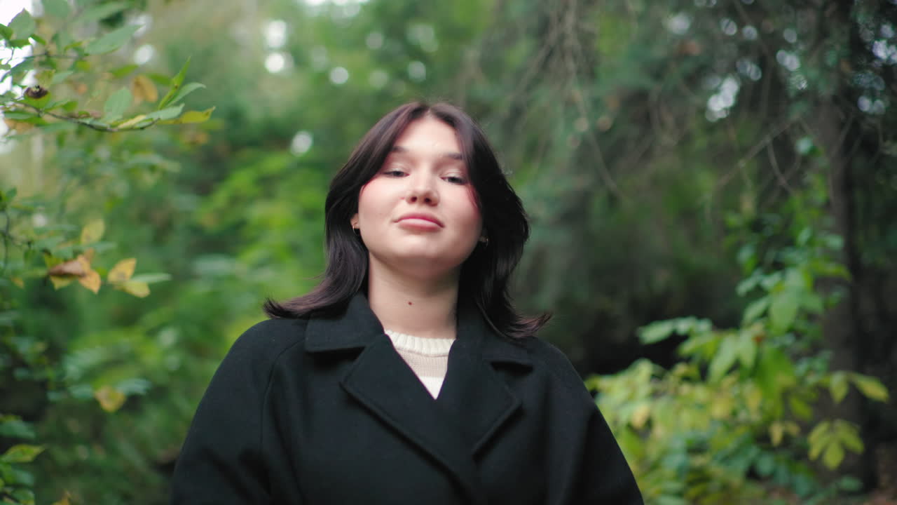 Portrait nature lover in black coat closes book and gazes around, standing among green forest foliage, calm expression, soft bokeh, peaceful autumn mood, mindful pause during leisurely outdoor walk