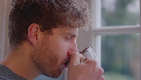 close up of young man at home thoughtfully looking out of window and drinking cup of coffee