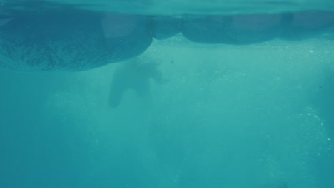 disparo submarino de la madre relajándose acostada en una cama de aire en la piscina mientras los niños saltan y la salpican