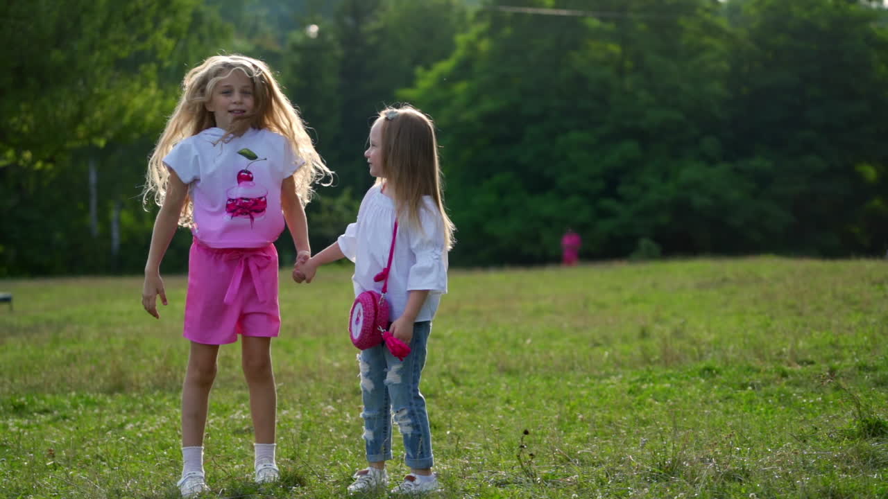Two small cute girls jump cheerfully outdoor in park. Happy friends outside hold hands while spending time on fresh air. Happy weekend concept.