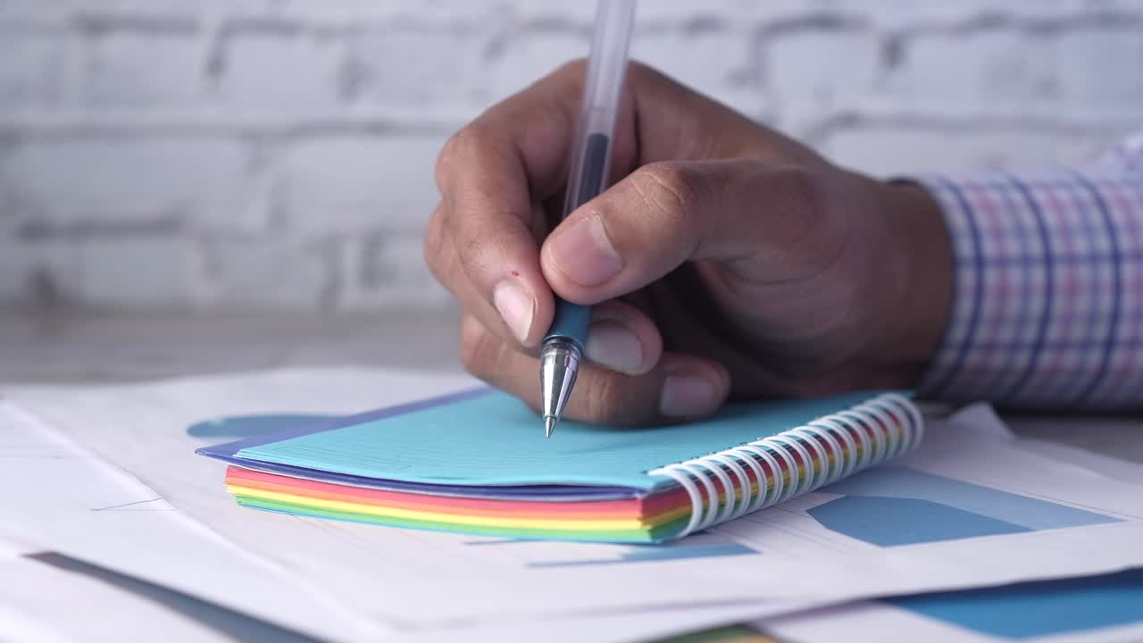 Person taking notes with a pen and spiral notebook on a desk with charts and graphs
