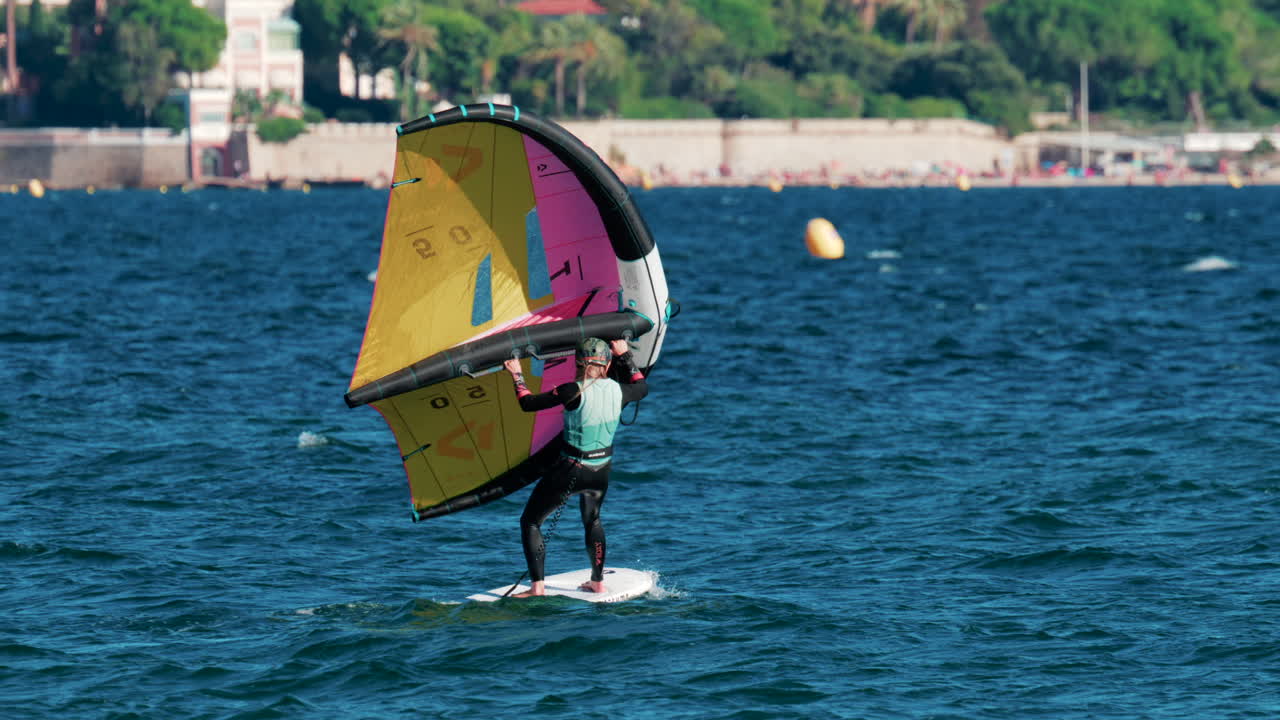 Cannes, France - October 6, 2025: A woman balances on a wing foil board holding a colorful yellow and pink sail on a sunny day
