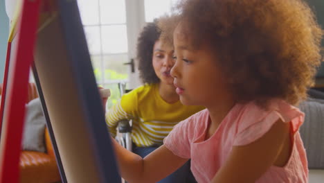 Madre-En-Silla-De-Ruedas-Viendo-A-Su-Hija-Divertirse-Dibujando-En-Una-Pizarra-En-Casa