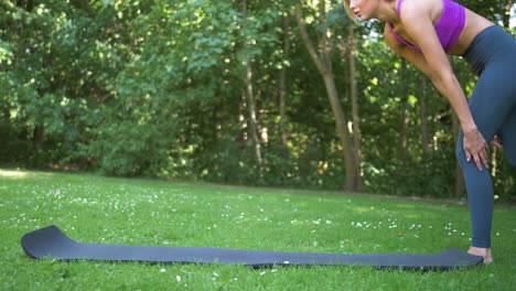 Young-woman-with-long-blonde-hair-unrolls-her-yoga-mat-in-a-park-before-sitting-and-meditating-in-her-pose