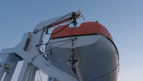 Vista-De-ángulo-Bajo-Del-Bote-Salvavidas-Colgado-En-La-Cubierta-Del-Ferry,-Cielo-De-Puesta-De-Sol,-Bote-Pequeño-De-Emergencia