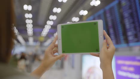 asian woman using white mobile tablet device with green screen in terminal hall while waiting her flight at the departure gate in international airport.