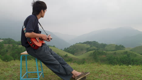 Solitary-emotional-young-man-playing-ukelele-on-tropical-hilltop-of-Southeast-Asia