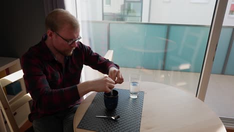 Man-Sits-at-Table,-Pours-Sugar-in-Cup-of-Coffee,-Stirs-with-Spoon