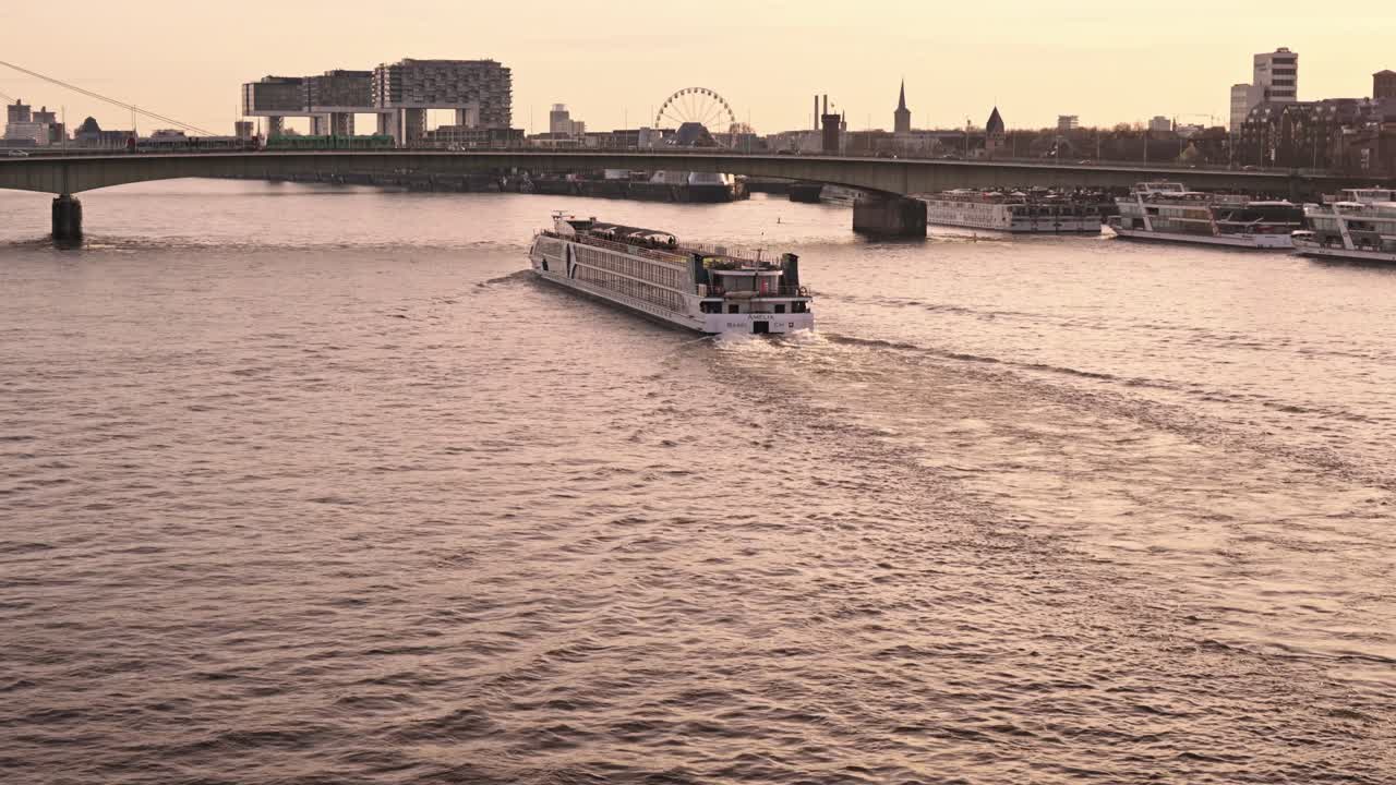 Et stort, hvitt elvecruiseskip som reiser langs Rhinen i Köln tatt under den gylne timen, med det varme, disige lyset fra den nedgående solen som kaster en myk glød over vannet