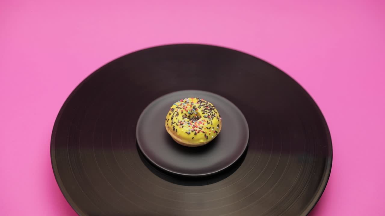 An American donut topped with yellow chocolate with black choco and colorful crumbs turns in a circle on a black turntable. The shot from above