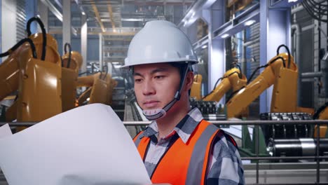 close up side view of asian male engineer with safety helmet looking at blueprint in his hands and looking around while standing with industrial robotic arms working