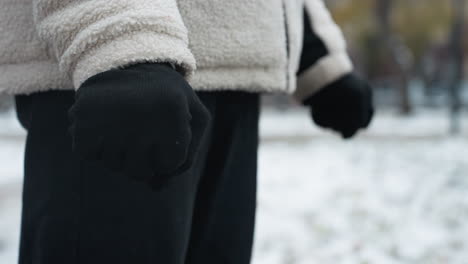 close-up side view of person wearing black gloves, winter jacket, and black trousers performing wrist twist, blurred snowy background with indistinct figures