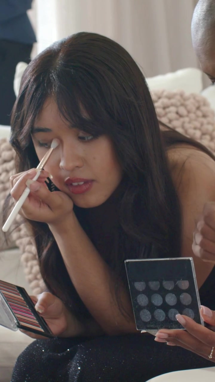 Vertical video: Opening palettes, two women applying eyeshadow on sofa using mirror, getting ready