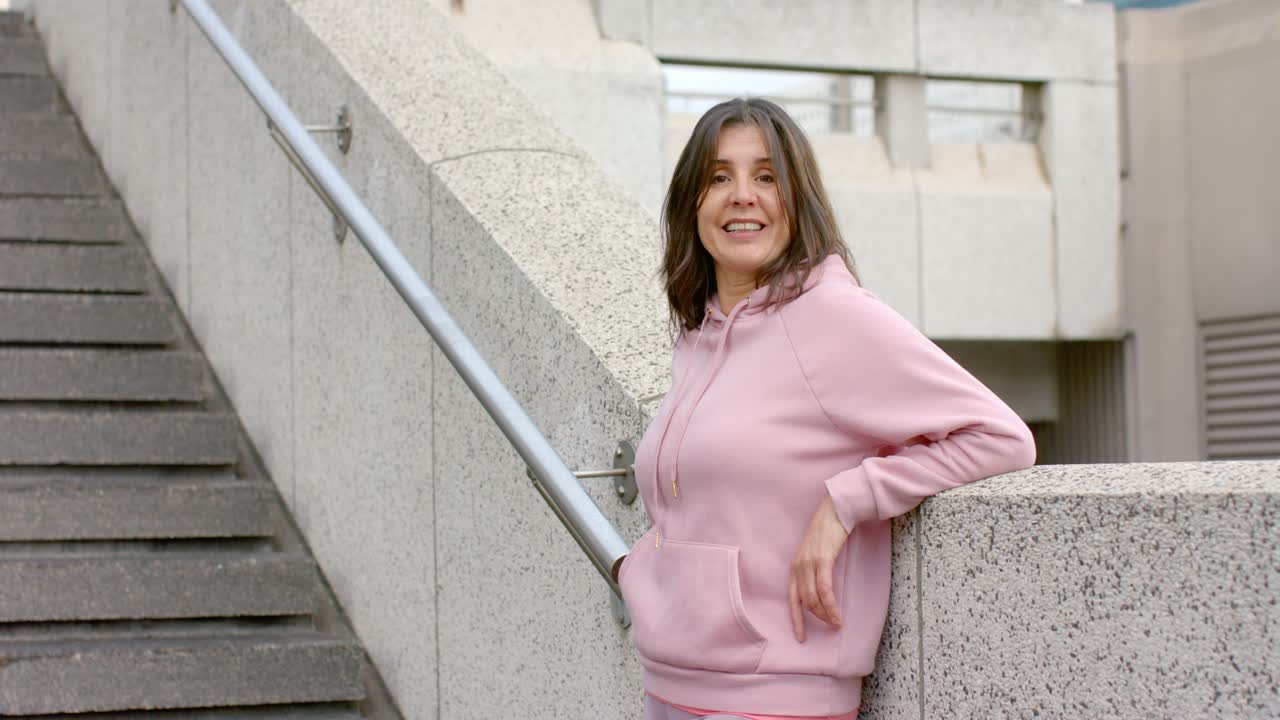 Donna reagisce a una panoramica della telecamera spostando il peso, girando lo sguardo e sorridendo accanto a una scala di cemento.