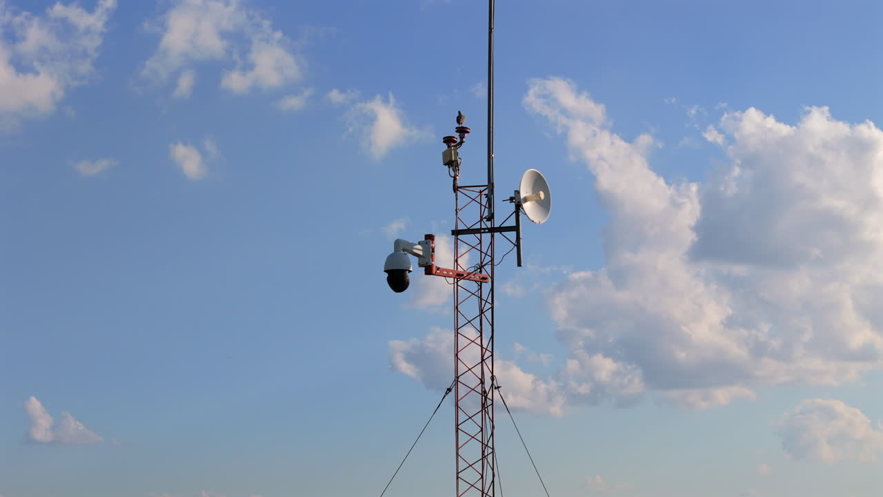 A bird standing at the top of a communications tower, set against a bright blue sky with clouds. featuring variety of antenna and surveillance equipment, including a dome camera, dolly in drone shot