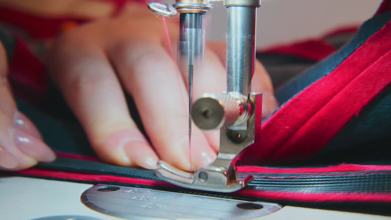 Close-up of hands guiding fabric under sewing machine needle. Thread pierces material with precision. Fabric shows red and dark blue stripes. Hands steady, focused on stitching task
