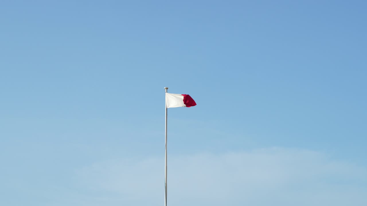 Maltese flag flying on a tall flag pole, waving in slow motion, with clear blue sky