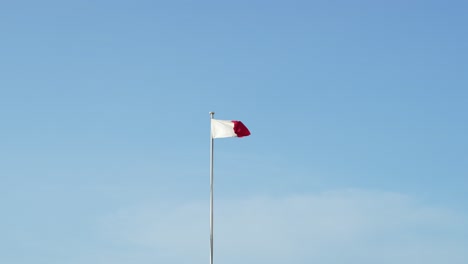 maltese flag flying on a tall flag pole, waving in slow motion, with clear blue sky