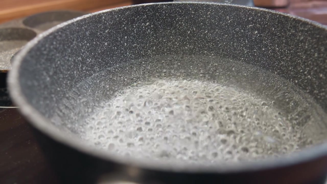 Low angled close view of speckled nonstick pan simmering on stovetop with rising bubbles, soft surface ripples, partial rim and handle visible, warm kitchen light, blurred background, and active