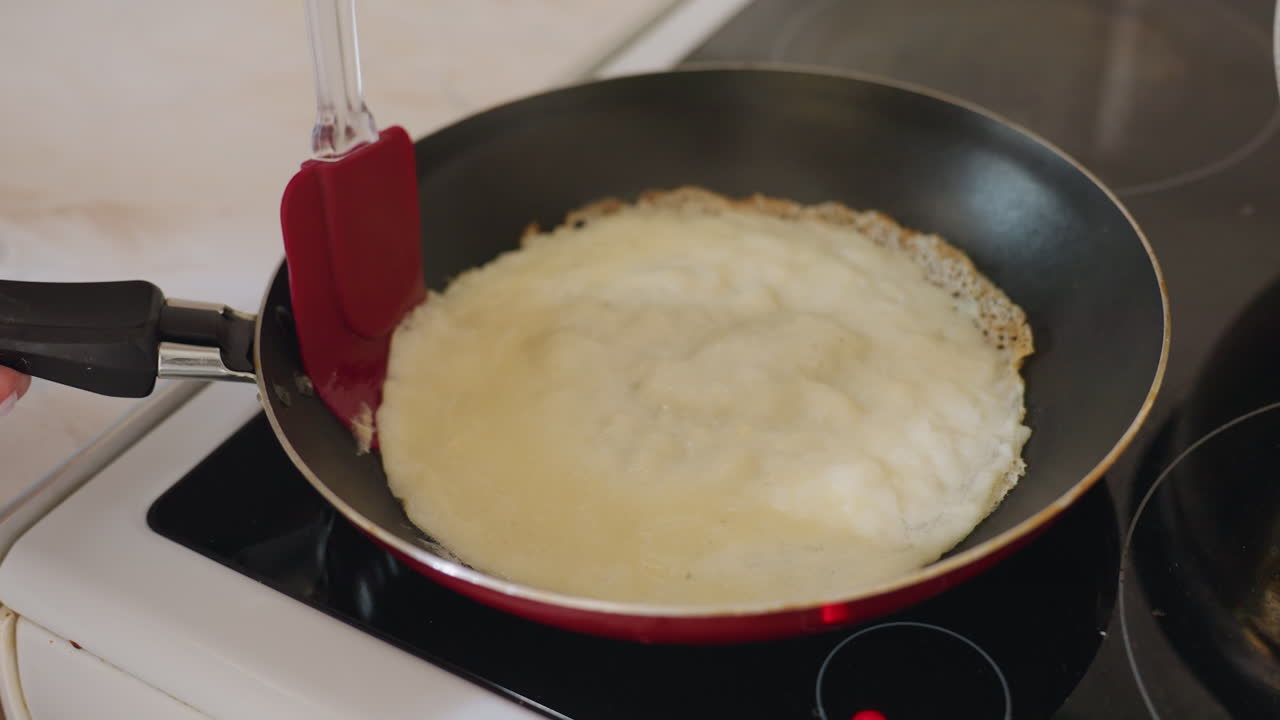 Close up of fried egg cooking in pan as someone carefully lifts it with red spatula over stove, showing texture and preparation process during breakfast meal in modern bright kitchen setting