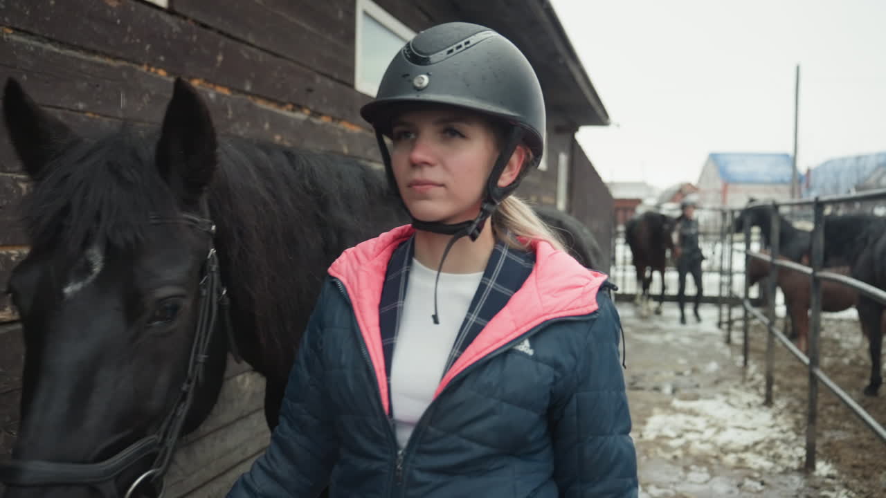 Kvinna leder svart sto längs stallet, ung ryttare i hjälm med rosa huvtröja håller tyglarna nära träladugårdsvägg, närbilder på sadel och pad, lerig gångväg och vinterkyla, nybörjarlektionskänsla.