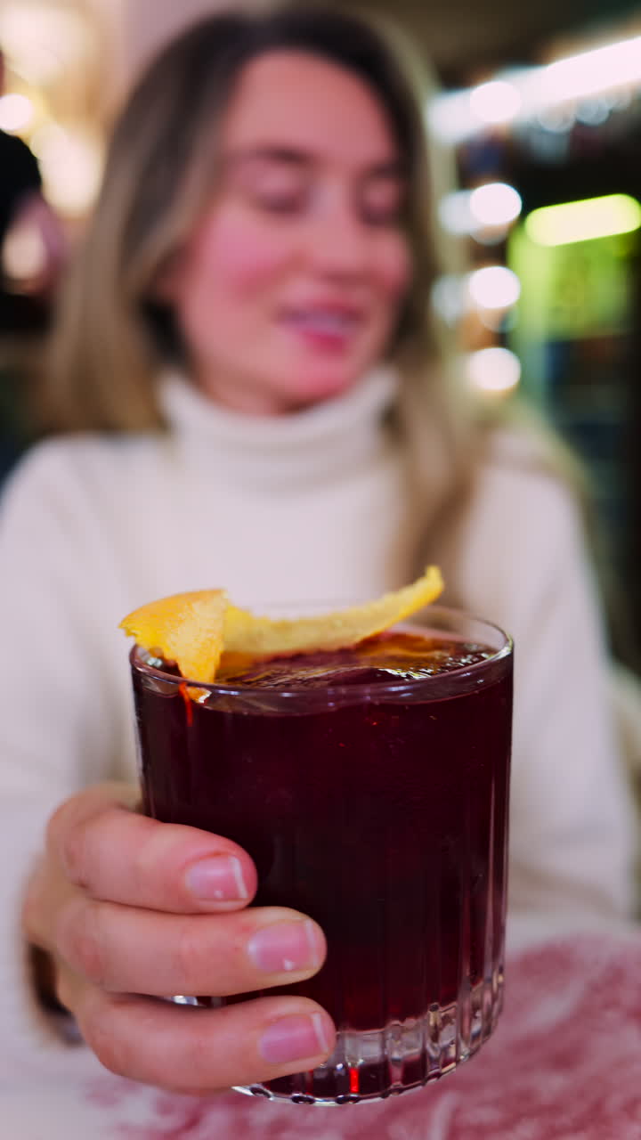 Close up of a woman holding a negroni cocktail on a red and white tablecloth at a restaurant. Vertical