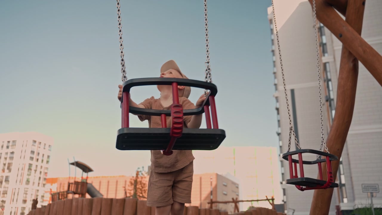 Un bambino si diverte sull'altalena rossa e nera in un parco giochi all'aperto. Indossa abiti marroni, inclusi pantaloncini e una maglietta, mentre si tiene alle catene per dondolarsi avanti e indietro.
