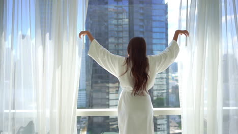 Mujer-Joven-En-Albornoz-Abriendo-Cortinas-De-Ventana-De-Dormitorio-En-Mañana-Soleada-Con-Hermosa-Vista-De-La-Ciudad-Y-Estirando-Las-Manos